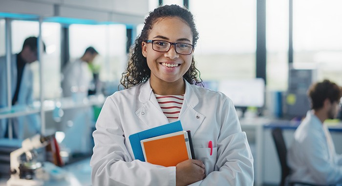 Person in white labcoat holding notebooks and smiling