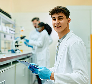 Man wearing white lab coat and surgical gloves holding a clipboard.